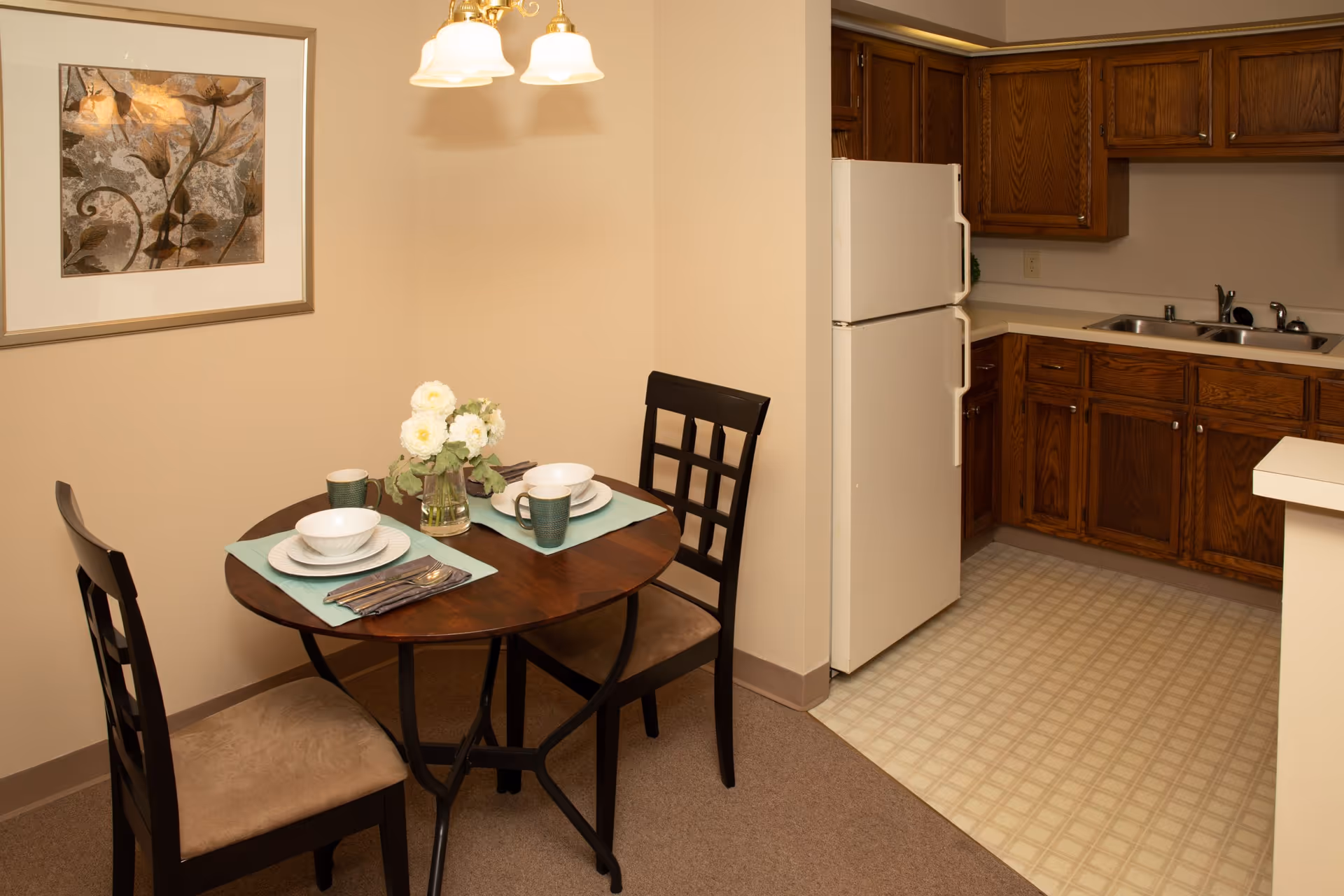 A small dining area with a round wooden table set for two, featuring white plates, bowls, green mugs, and silverware on light blue placemats. A vase with white flowers is in the center of the table. Two wooden chairs with cushioned seats are placed around the table. Adjacent to the dining area is a kitchen with wooden cabinets, a white refrigerator, a double sink, and a light-colored tiled floor.
