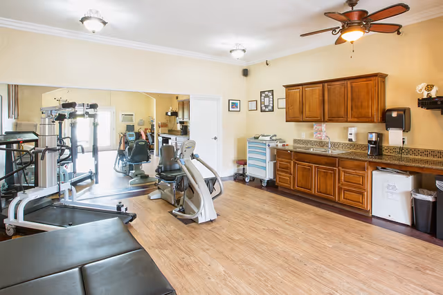 Bright interior room with exercise equipment, wood cabinets and a sink under a ceiling fan.