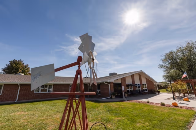 Outdoor view of a nursing and rehabilitation center building with a red and white windmill sculpture in the foreground, green grass, a clear blue sky with the sun shining brightly, and some trees and pumpkins near a patio area with an American flag.