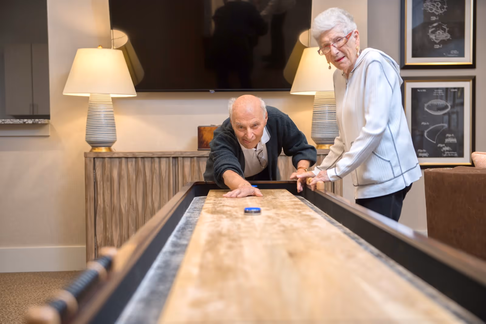 An elderly man and woman playing shuffleboard indoors in a well-lit room with two table lamps, a large flat-screen TV mounted on the wall, and framed artwork in the background.