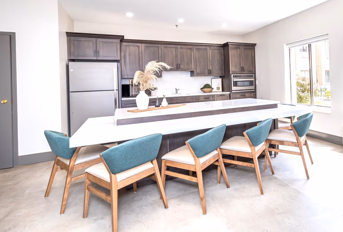 Bright modern kitchen with dark wooden cabinets, a stainless steel refrigerator, built-in microwave and oven, a white island countertop with decorative vases, and six wooden chairs with teal backs and beige seats arranged around the island.