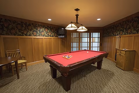 A cozy game room with a red felt pool table in the center, surrounded by wood-paneled walls and floral wallpaper above. The room features a hanging light fixture above the pool table, a wooden chair and small round table on the left, and a wooden cabinet with pool cues on the right. French doors with frosted glass panels are visible at the back of the room.