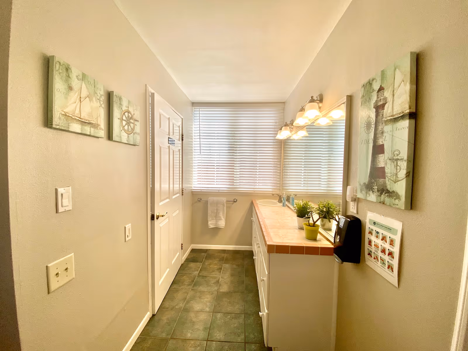 Bright bathroom with a long vanity and mirror, countertop plants, towel rack, tiled floor, and nautical wall art.
