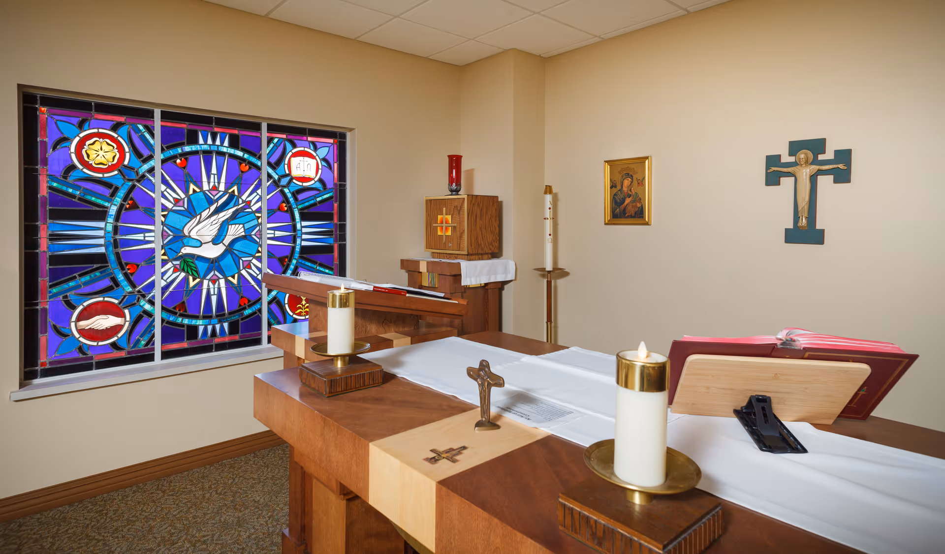 Small chapel interior with a wooden altar, lit candles, a crucifix, and a colorful stained-glass window depicting a dove.