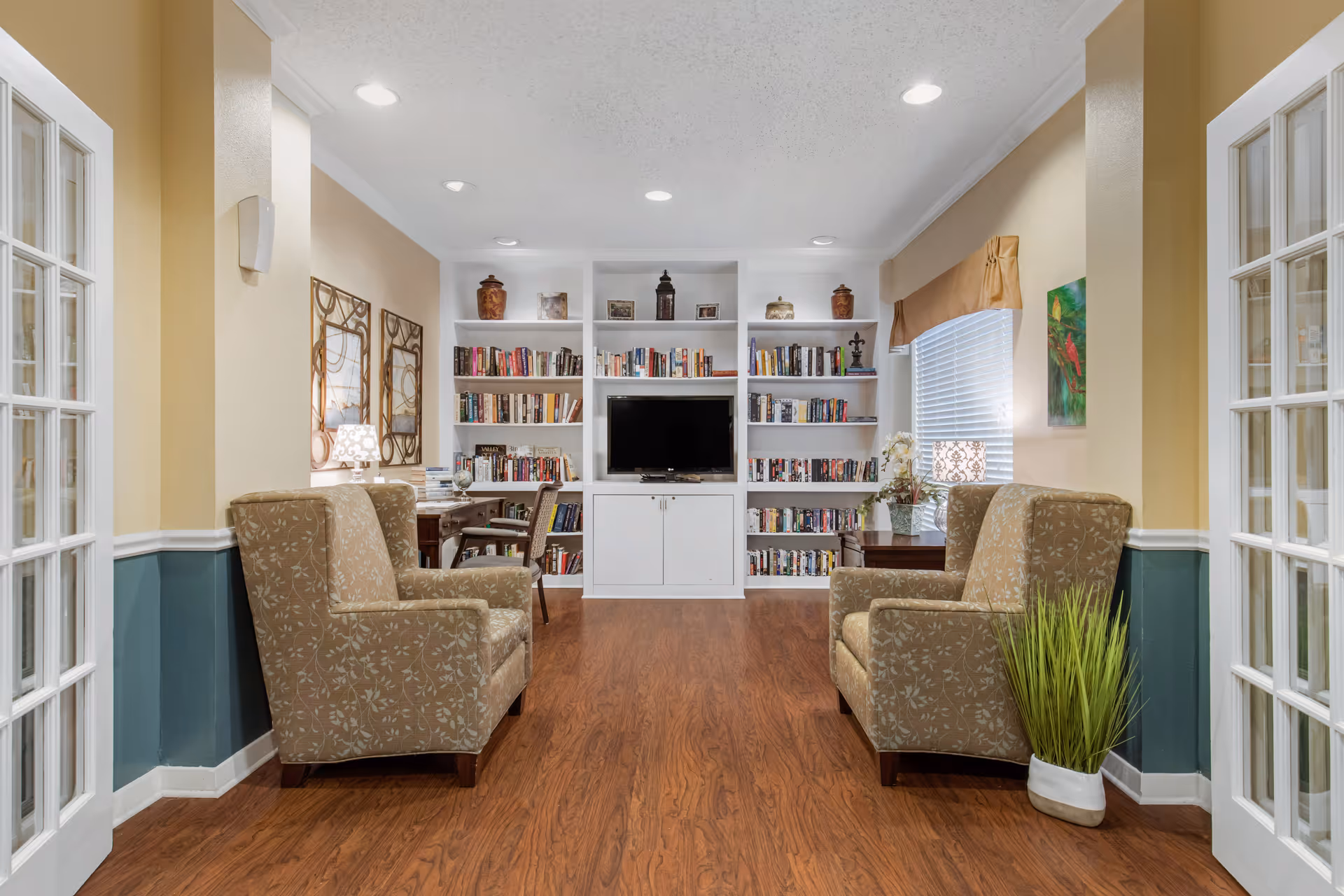 Cozy sitting area with upholstered armchairs, built-in bookshelves and a central TV in a bright common room.