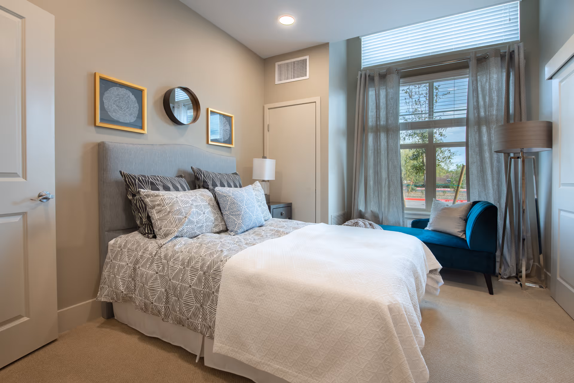A cozy bedroom with a neatly made bed featuring patterned pillows and a white blanket. Above the bed are two framed artworks and a round mirror. To the right, there is a large window with sheer curtains, a teal upholstered chair with a pillow, and a tall floor lamp. The room has beige walls and carpeted flooring.