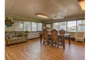 Open common room with wood floors featuring a dining table and chairs, a couch, large windows with blinds, and a ceiling fan.
