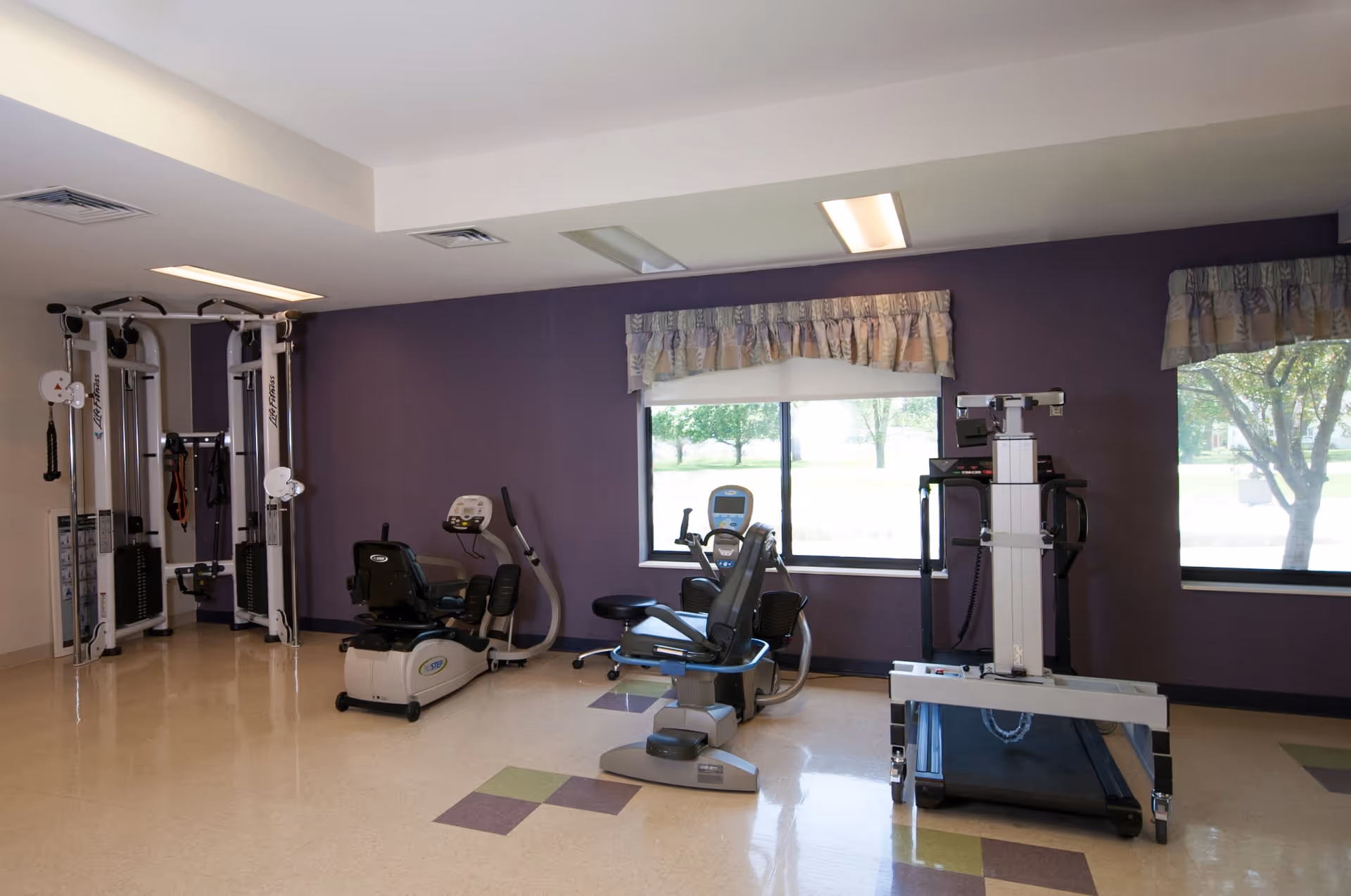 A fitness room with exercise equipment including a recumbent bike, a treadmill, and a multi-function weight machine. The room has a purple accent wall with two windows covered by valances, allowing natural light to enter. The floor has a tiled pattern with purple and green squares.