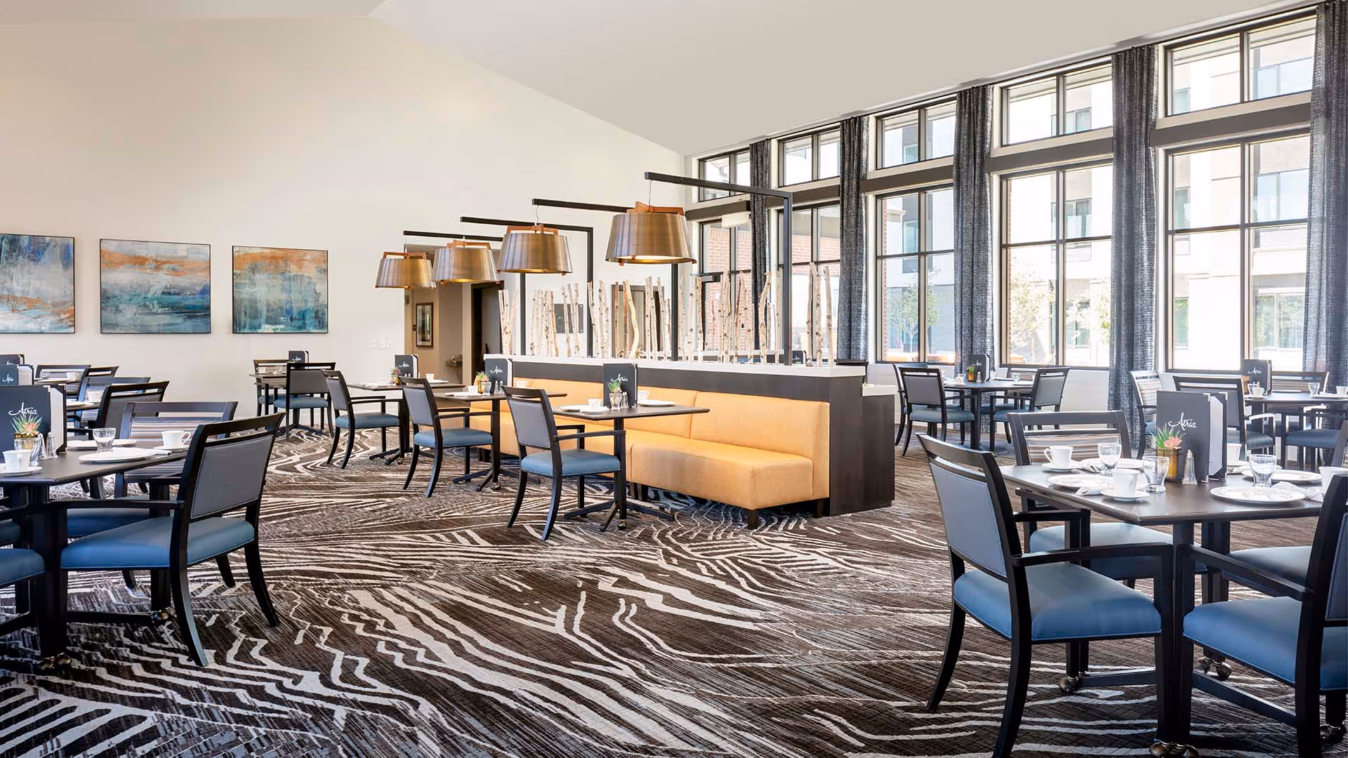 Bright modern dining room with multiple tables and chairs, a central banquette with pendant lights, and large floor-to-ceiling windows.