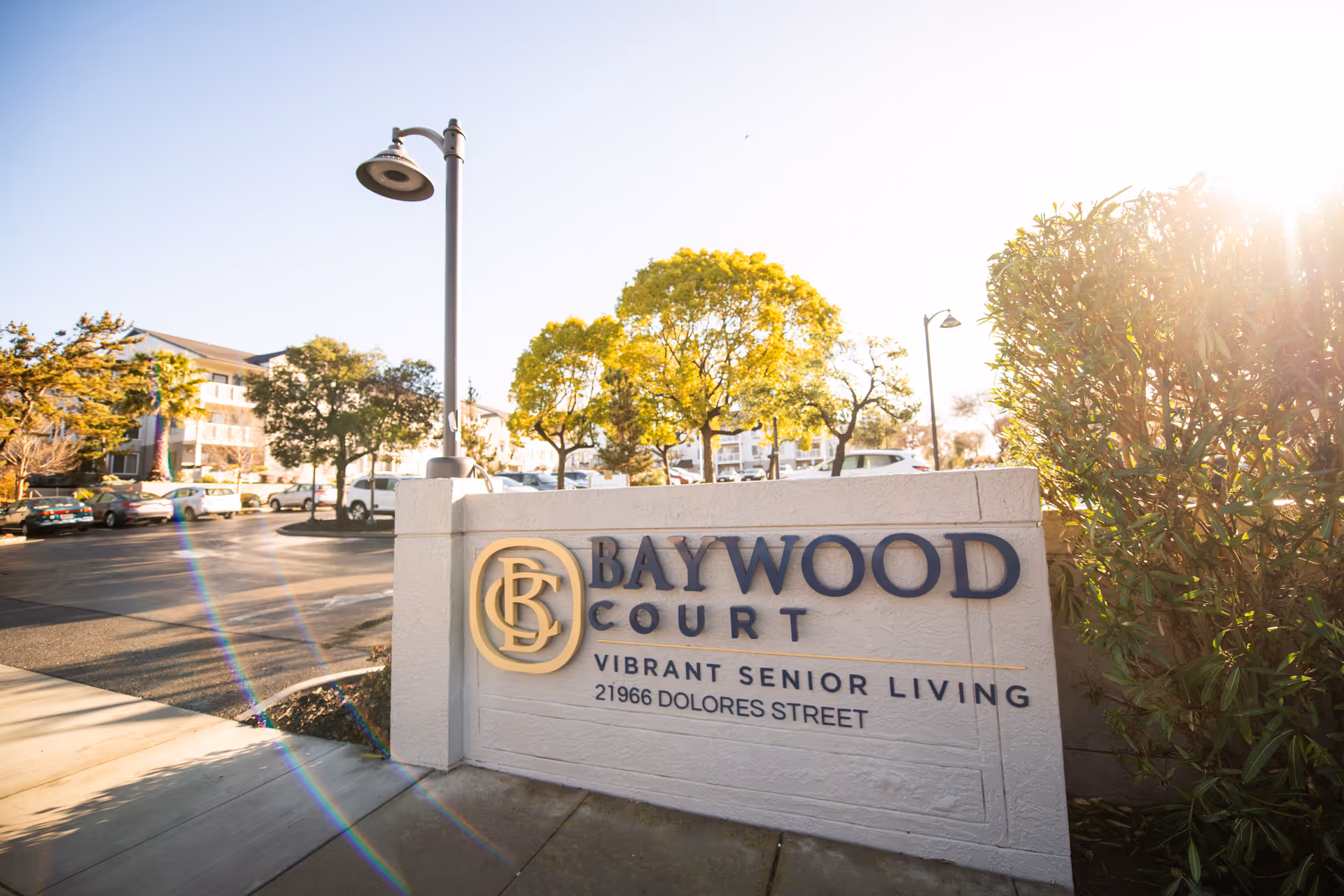 Outdoor view of the entrance sign for Baywood Court, a senior living facility, with trees, parked cars, and a building in the background under a clear sky with sunlight.