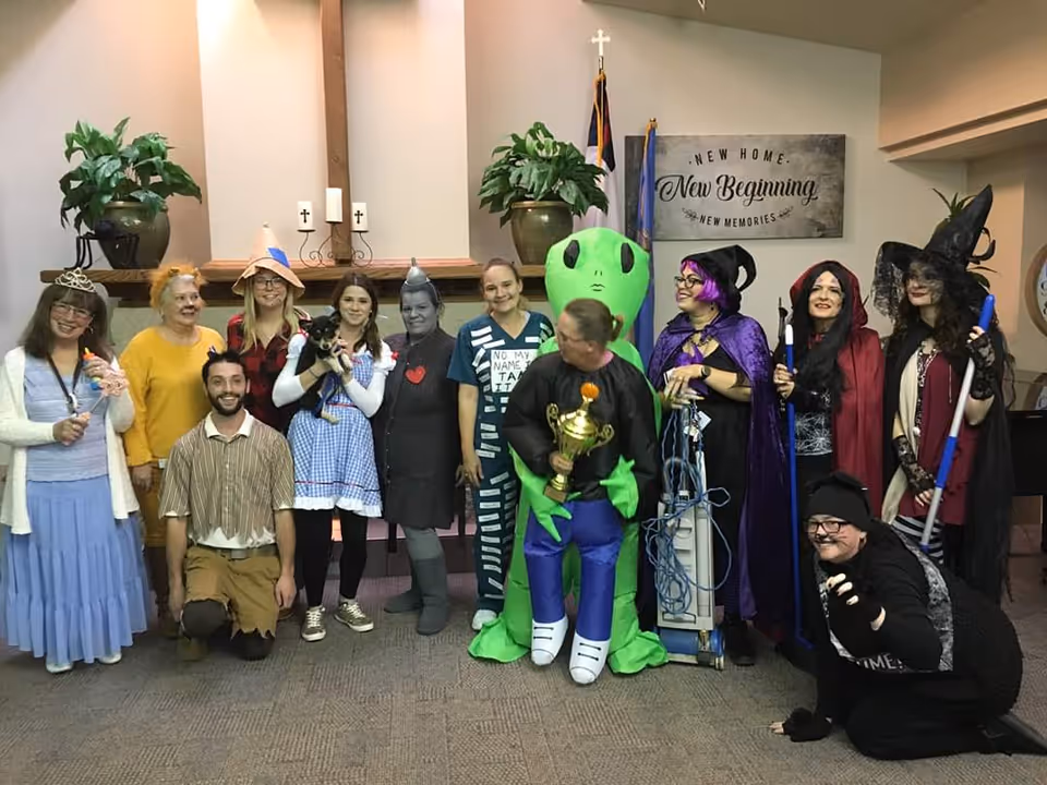 A group of adults in Halloween costumes, including an inflatable alien and several witches, posing together in a decorated community room with a 'New Beginning' sign on the wall.