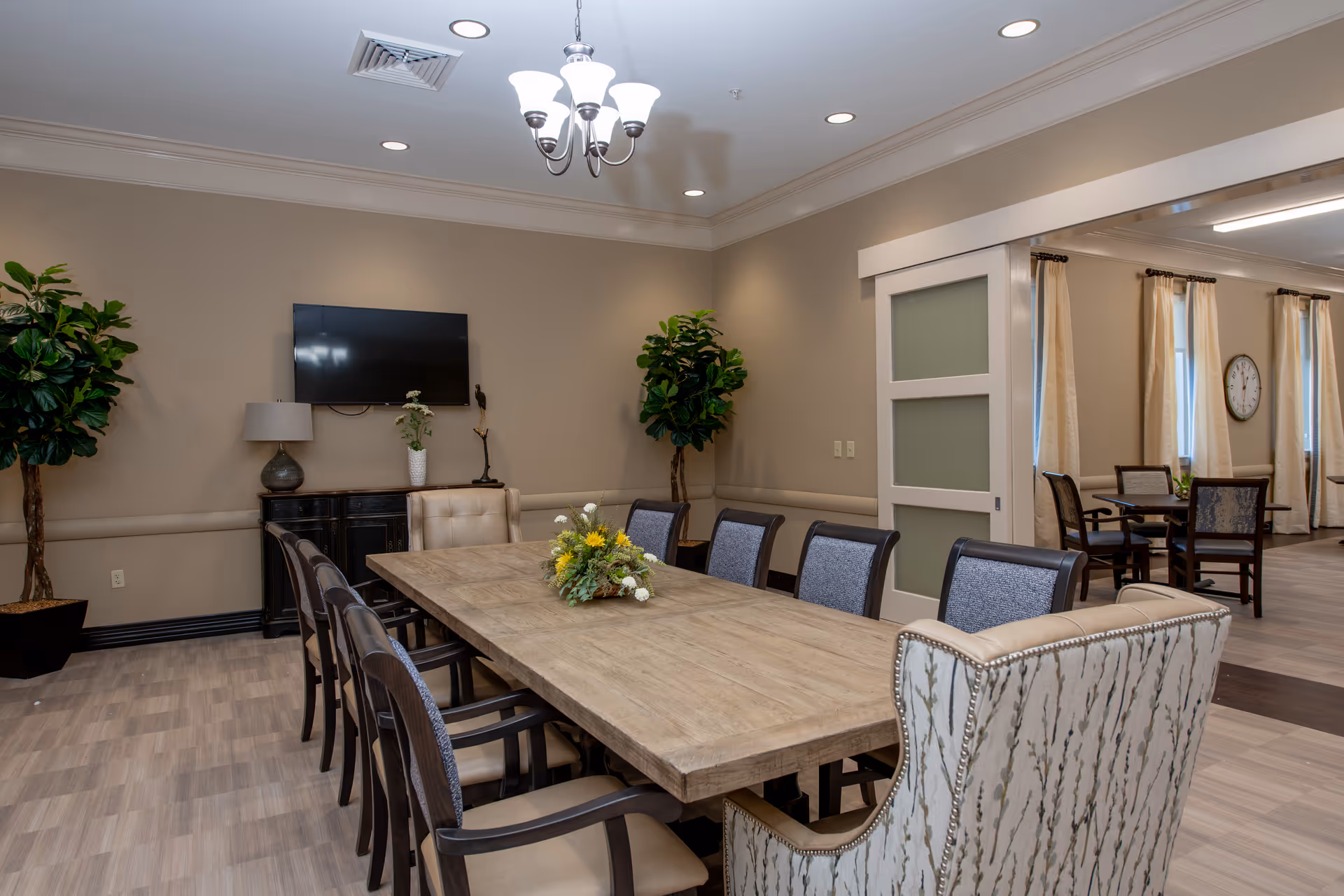 A well-lit dining room in an assisted living facility featuring a long wooden table surrounded by cushioned chairs. The room has beige walls, two large potted plants, a wall-mounted flat-screen TV, and a sideboard with decorative items. A chandelier hangs from the ceiling, and an adjacent room with additional tables and chairs is visible through a wide doorway.
