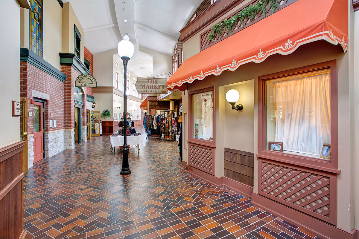 Indoor hallway designed to resemble a small town street with storefronts including a cafe and a market. The floor is tiled with a brick pattern, and there are streetlamp-style lights along the corridor. The walls have a mix of brick and wood paneling with decorative windows and awnings.
