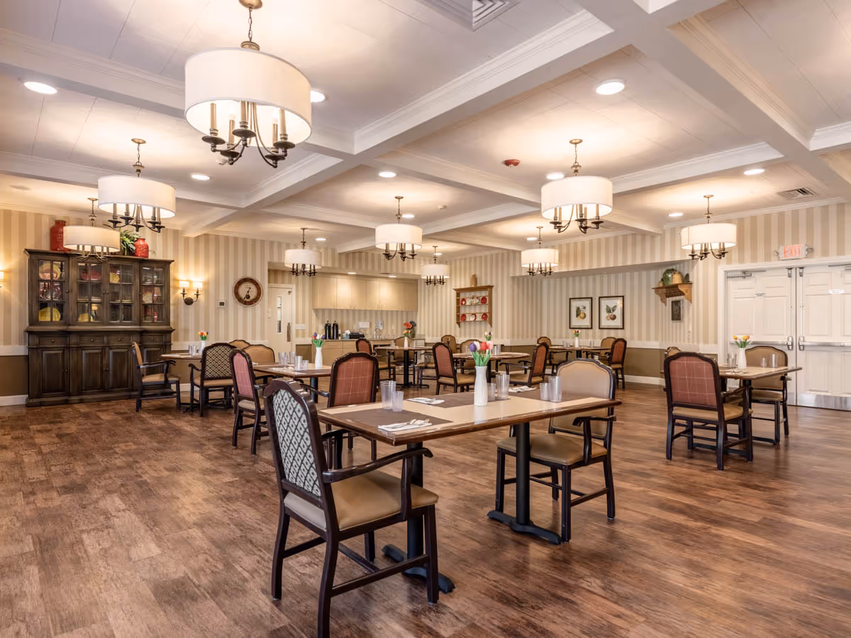 A spacious dining room with multiple tables and chairs arranged neatly. Each table has a vase with flowers and place settings. The room features wooden flooring, beige striped wallpaper, and multiple ceiling lights with drum shades. There is a wooden cabinet with glass doors on the left side and framed artwork on the walls.