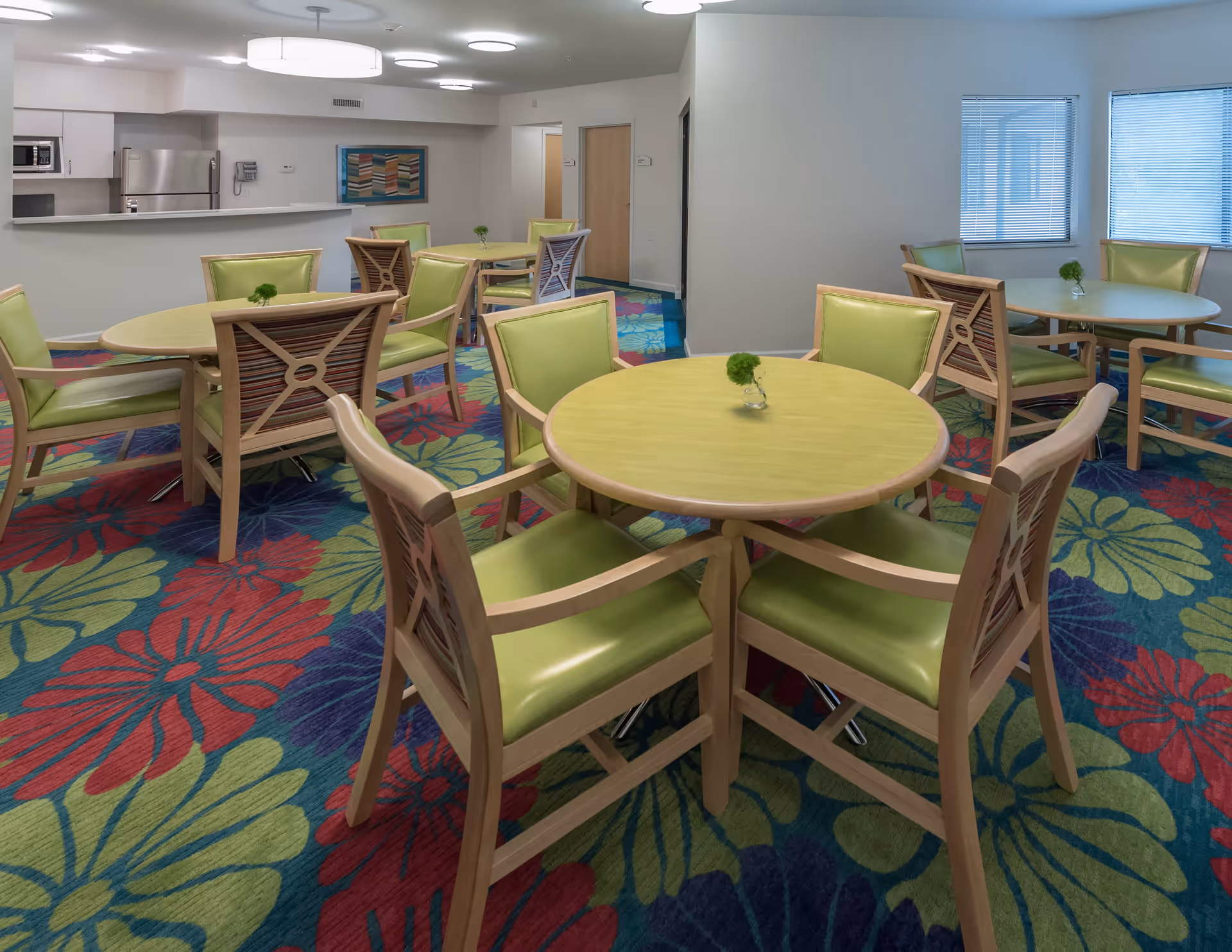 A bright and clean dining area with round wooden tables and green cushioned chairs arranged on a colorful floral carpet. The room has large windows with blinds, a kitchen area with stainless steel appliances, and neutral-colored walls with a framed artwork.
