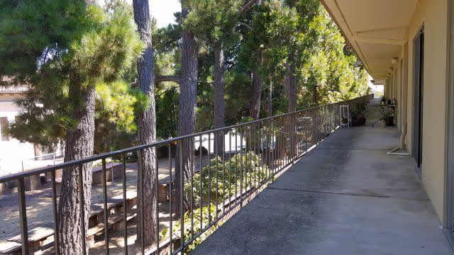 A long outdoor corridor with a metal railing on the left side overlooking a garden area with tall pine trees and picnic tables. The corridor is adjacent to a building with doors and windows on the right side under a covered walkway.
