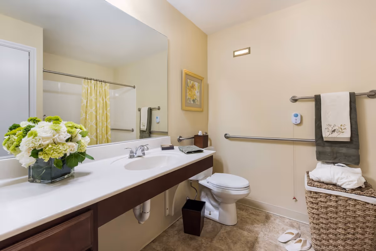 A clean and well-lit bathroom featuring a white sink with a large mirror above it, a toilet with grab bars on the wall, a wicker laundry basket with towels, a pair of white slippers on the floor, and a yellow patterned shower curtain. There is also a vase with green and white flowers on the sink counter and a framed floral picture on the wall.