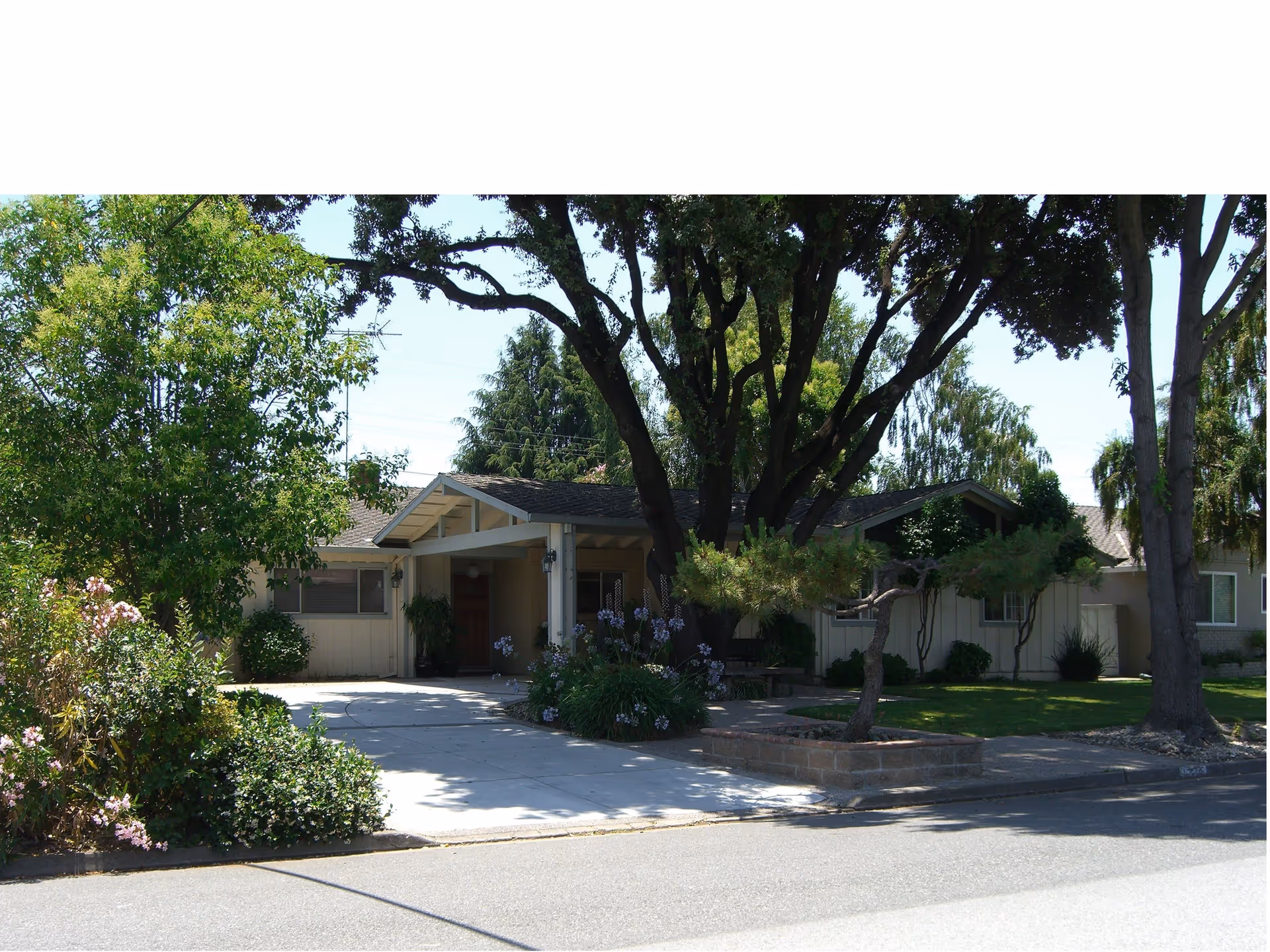 Exterior view of a single-story residential care home with a driveway, surrounded by trees and shrubs under a clear sky.