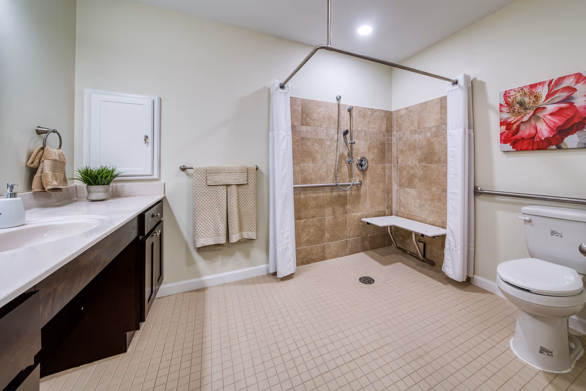 A spacious senior living bathroom with beige tiled floor and walls in the shower area. The shower has a fold-down seat, grab bars, and a handheld showerhead with a white curtain on a ceiling-mounted rod. There is a white toilet with grab bars on the right side and a dark wood vanity with a white countertop and sink on the left. Beige towels hang on a towel rack and ring, and a potted plant and soap dispenser are on the vanity. A large red and white flower painting is on the wall above the toilet.