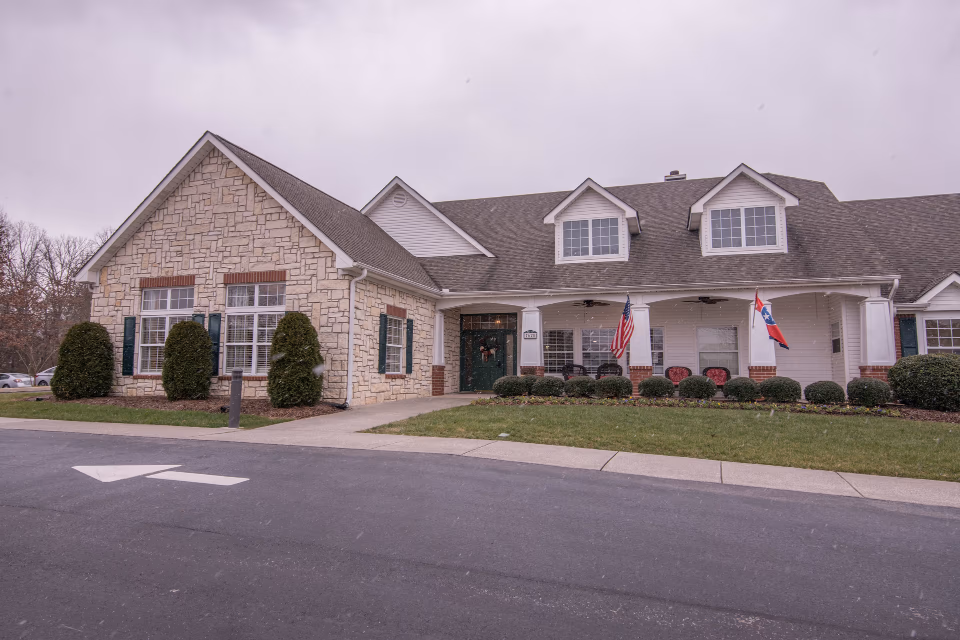 Exterior view of a single-story senior living facility building with stone and white siding, a covered porch with chairs, American and Tennessee state flags, and neatly trimmed bushes and lawn.