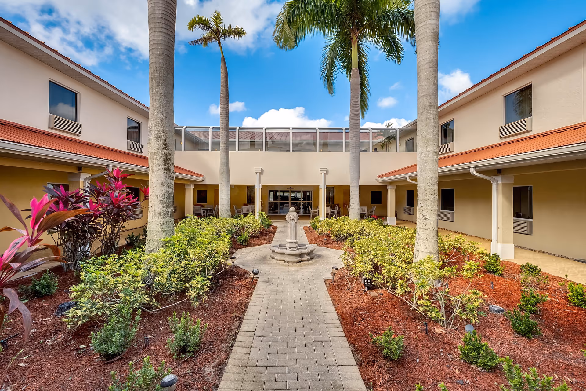 Outdoor courtyard area of Balmoral Assisted Living featuring a paved walkway leading to a central fountain, surrounded by landscaped garden beds with shrubs and palm trees. The courtyard is enclosed by a two-story building with beige walls and red roofs under a partly cloudy blue sky.