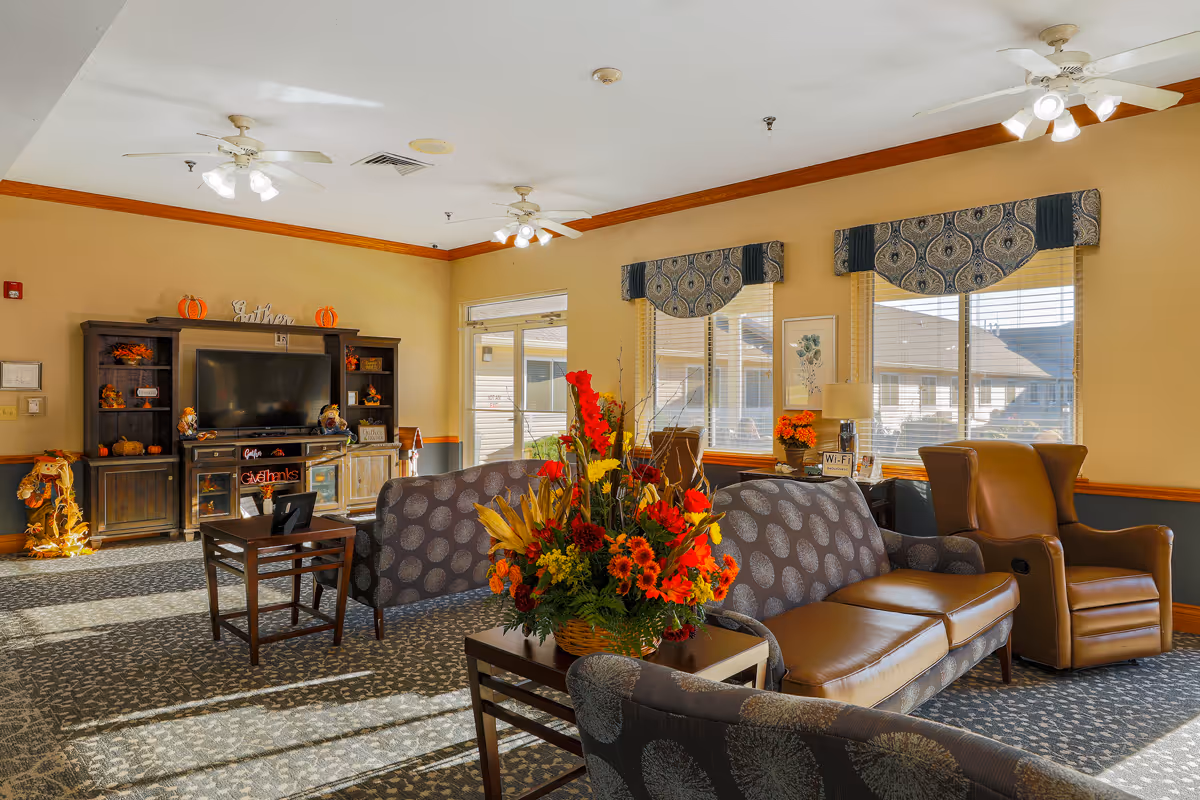 A cozy living room area in an assisted living facility with patterned sofas, a leather armchair, and a coffee table with a large floral arrangement. The room has large windows with decorative valances, ceiling fans with lights, and a wooden entertainment center with a TV and autumn-themed decorations.
