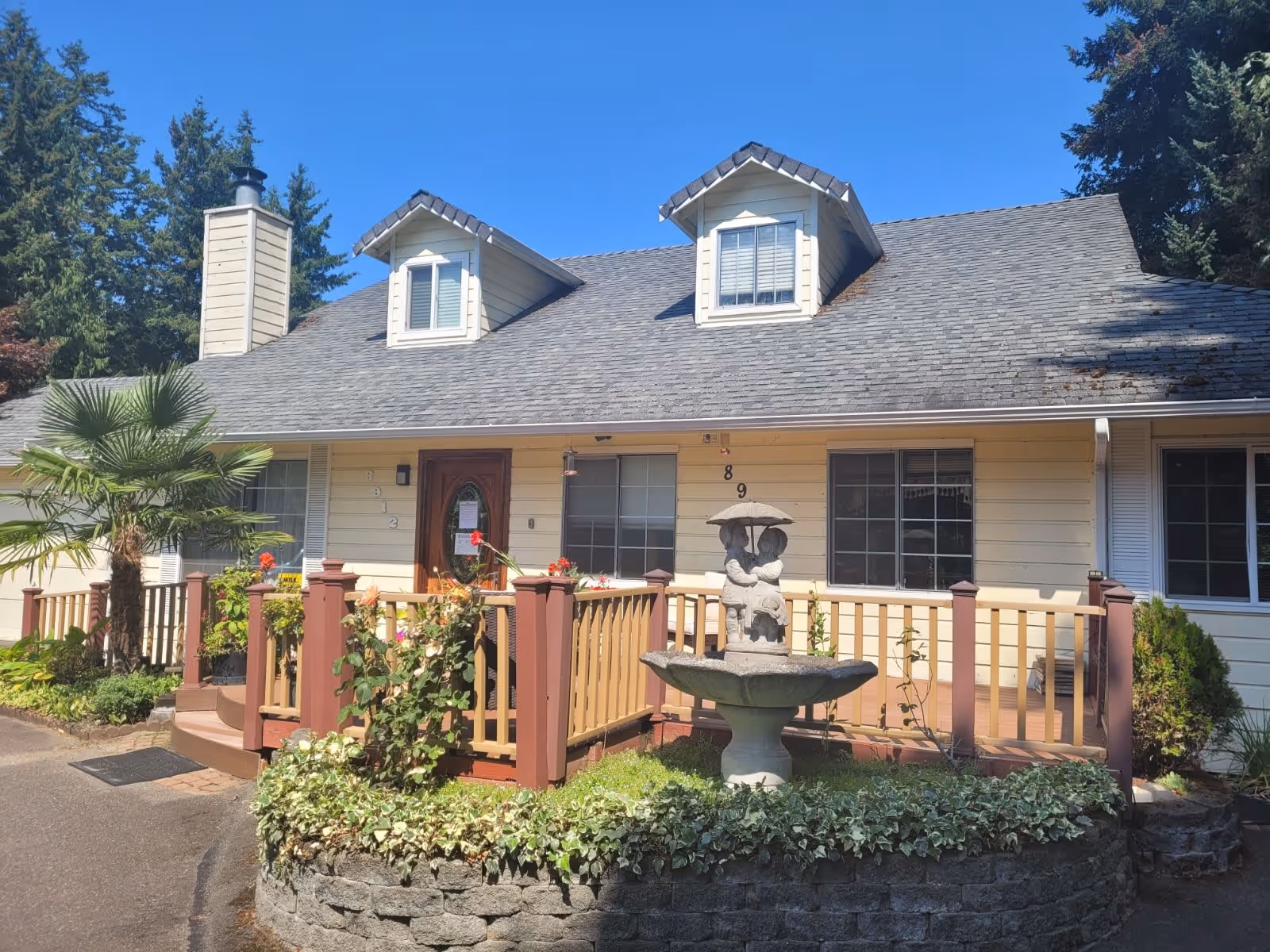 Exterior view of a single-story residential building with beige siding and a gray shingled roof featuring two dormer windows. The front porch has a wooden railing with red and beige colors, decorated with plants and flowers. In front of the porch, there is a stone fountain with a sculpture of two children holding an umbrella. The area is surrounded by greenery and trees under a clear blue sky.
