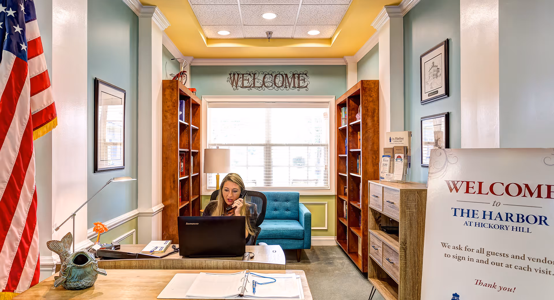 Reception area with a staff member on the phone behind a desk, flanked by bookshelves, an American flag, a 'Welcome' sign, and seating.