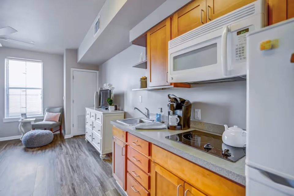 Studio-style kitchen with wooden cabinets, microwave, stovetop and sink opening into a small seating area with a chair and window.