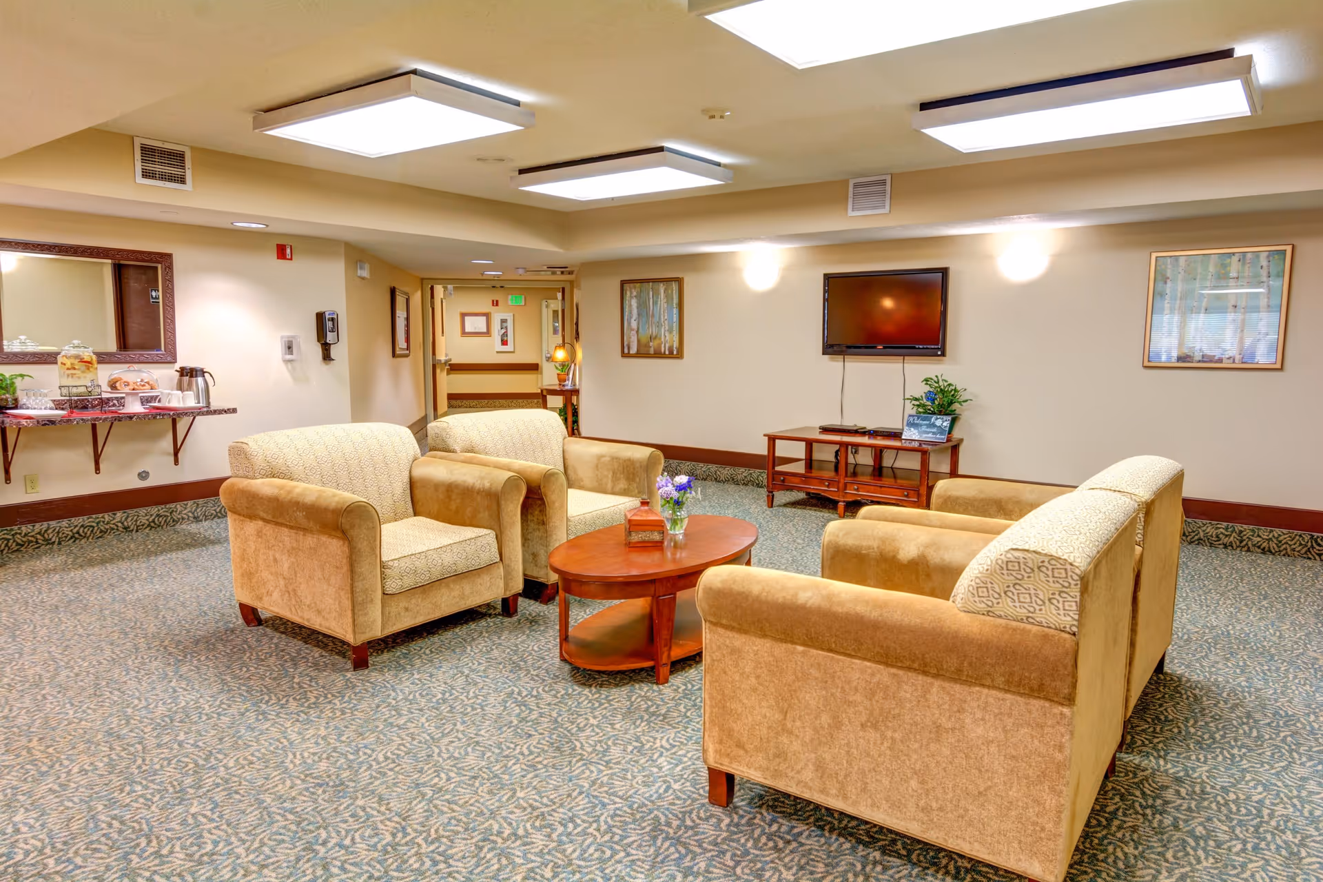 A cozy senior living facility common area with four beige armchairs arranged around a wooden coffee table with a small flower vase. The room has patterned carpet, a wall-mounted flat screen TV, framed artwork on the walls, and a refreshment station with drinks and snacks on the left side.