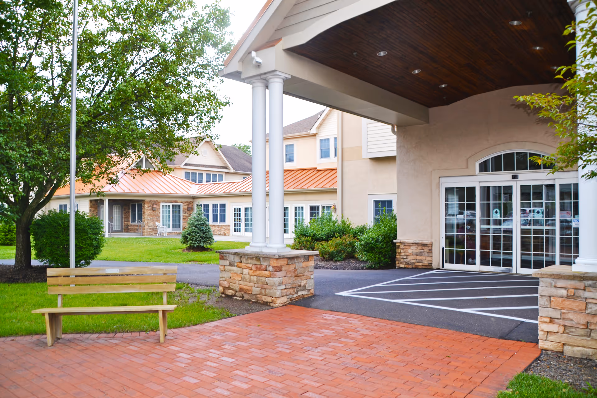 Exterior view of a senior living facility entrance with brick walkway and bench.