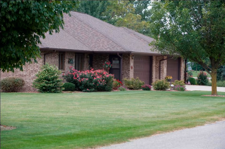 Exterior view of a single-story brick building with a well-maintained lawn, shrubs, and trees in front. The building has a sloped roof and two garage doors visible, with a driveway on the right side.