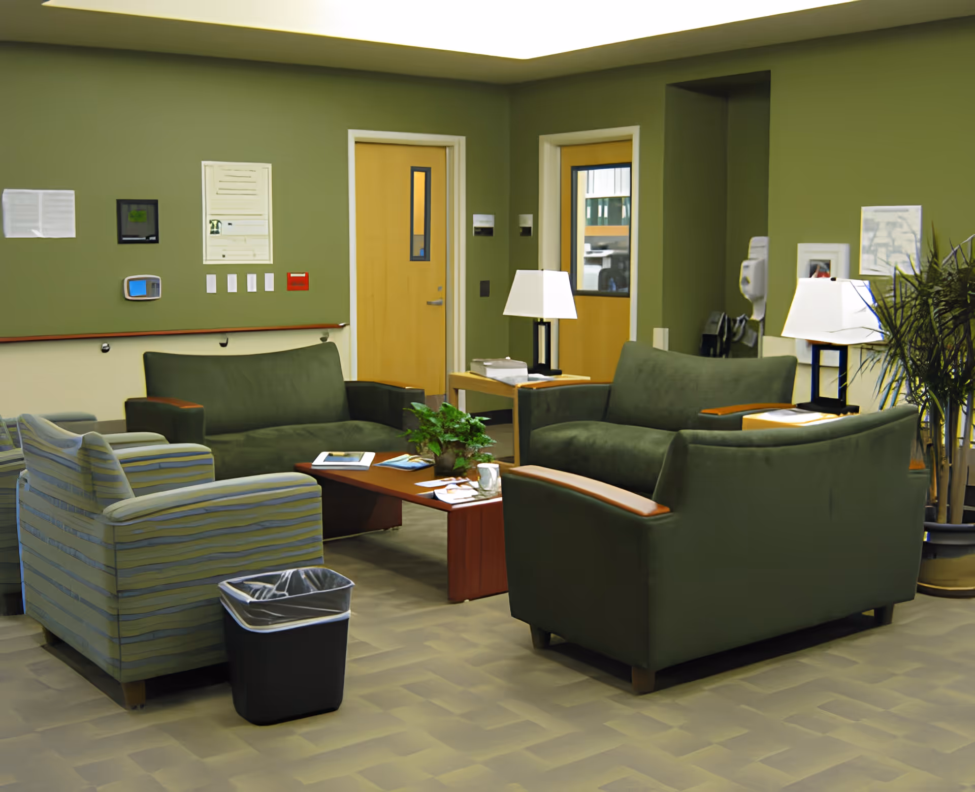 A communal lounge with green sofas and armchairs arranged around a coffee table, lamps, and potted plants against olive-green walls.