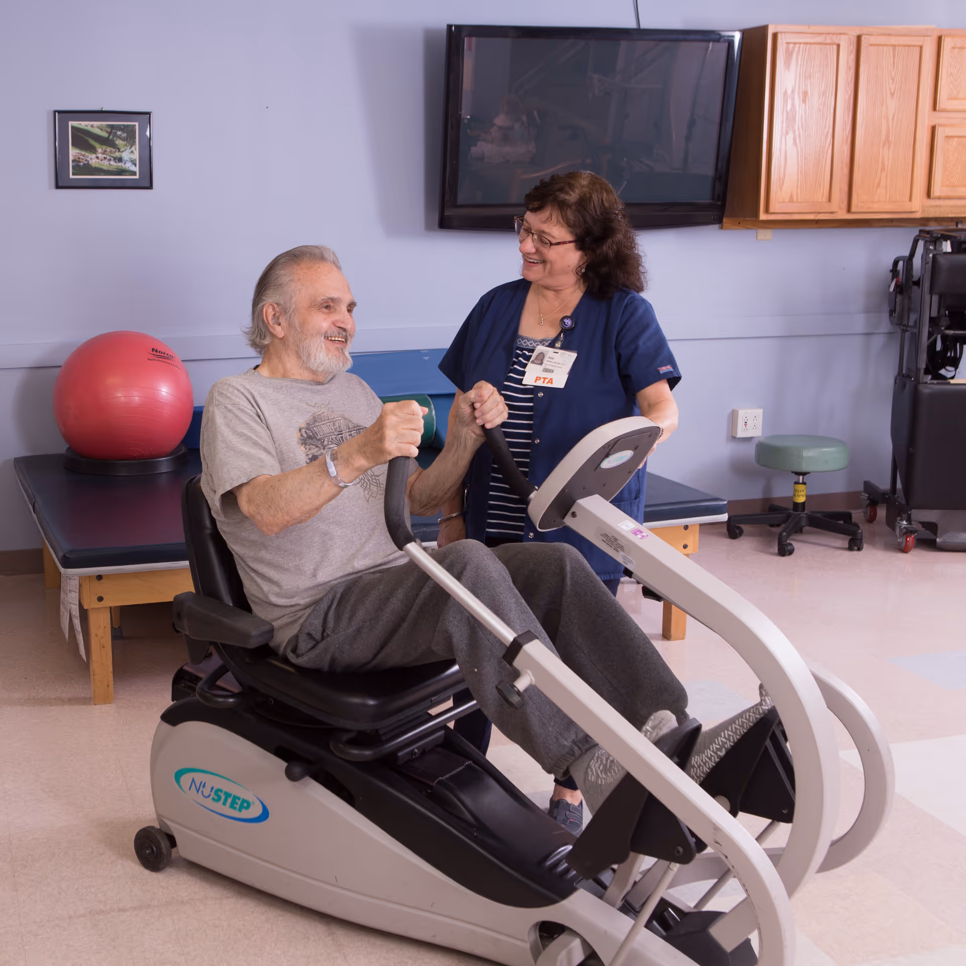 An elderly man is exercising on a NuStep recumbent cross trainer machine while a female physical therapist in navy scrubs smiles and supports him in a rehabilitation room with a red exercise ball, a bench, and a wall-mounted TV.