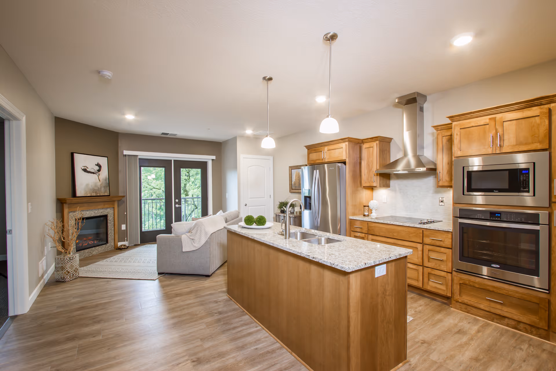 Open-concept kitchen with a central island, wooden cabinets and stainless steel appliances opening to a living area with a couch, fireplace and balcony doors.