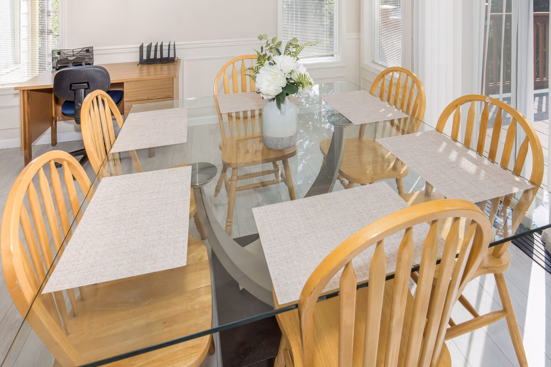 Glass-top dining table with six wooden chairs, placemats and a vase of flowers in a bright dining area with a small desk in the background.