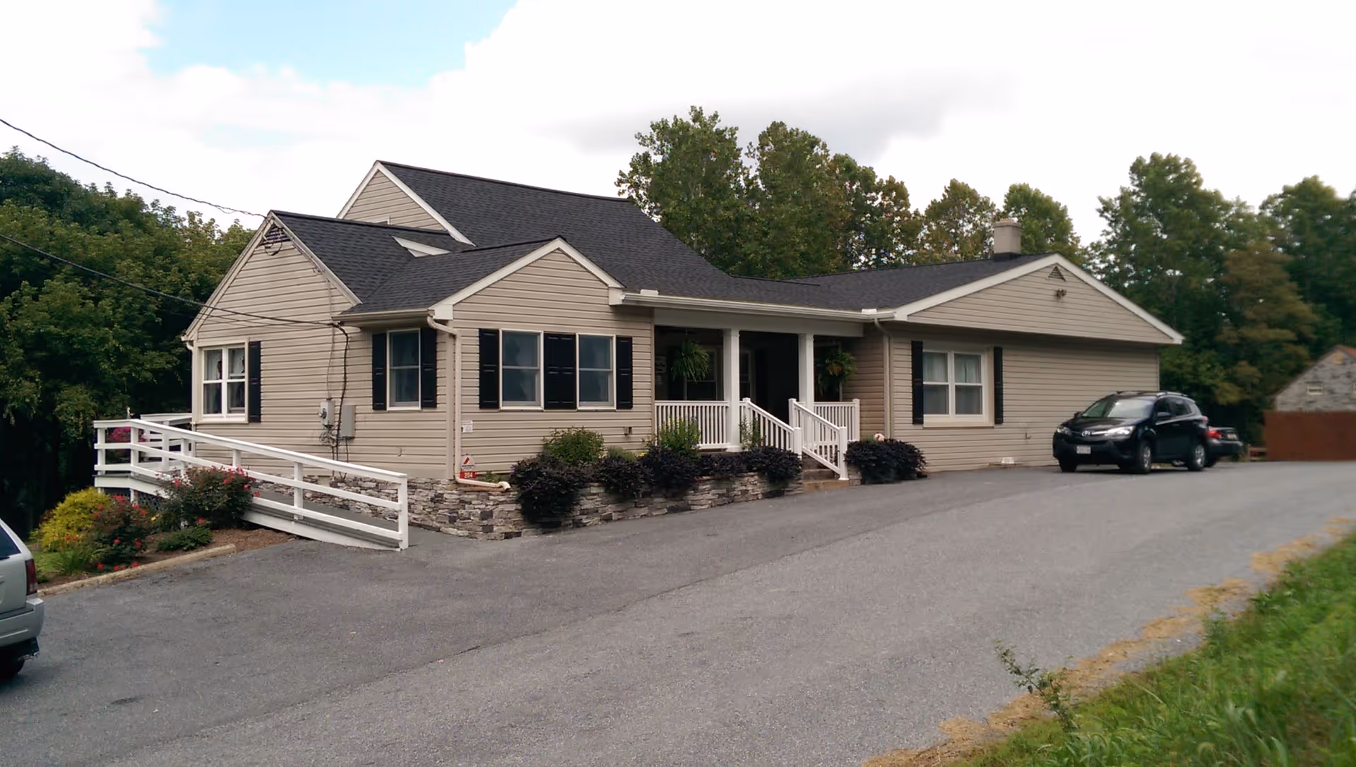 Exterior view of a single-story beige house with black shutters and a black roof. The house has a white railing along the front porch and a wheelchair ramp on the left side. There are bushes and plants along the front of the house, and two cars are parked on the driveway. Trees are visible in the background under a partly cloudy sky.