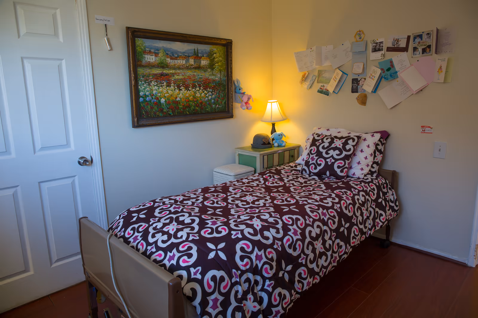 A single bed with a patterned purple, white, and pink bedspread in a small bedroom. There is a nightstand with a lamp, a hat, and a stuffed animal next to the bed. Above the nightstand, a bulletin board is covered with various cards and photos. A framed painting of a landscape with flowers hangs on the wall above the bed. A closed white door is visible on the left side of the room.