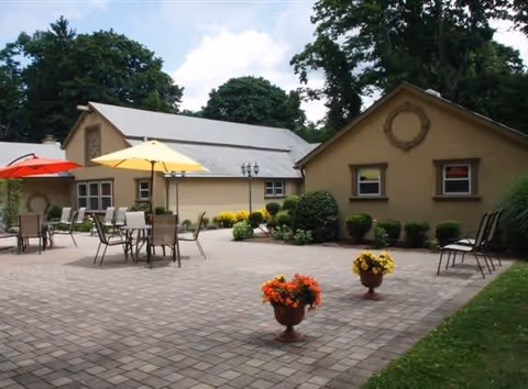 Outdoor patio area with several tables and chairs, some shaded by red and yellow umbrellas. The patio is paved with bricks and decorated with potted flowers. Surrounding the patio are beige buildings with windows and some greenery, including bushes and trees in the background under a partly cloudy sky.