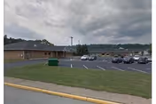 Exterior view of a single-story brick building with a parking lot in front. Several cars are parked in the lot, and there is a green dumpster on the grass near the building. The sky is cloudy.