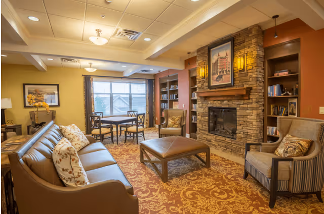 A cozy living room with a brown leather sofa adorned with floral pillows, two armchairs, a large ottoman coffee table, a stone fireplace with a framed picture above it, built-in bookshelves on either side, a round table with four chairs near a window with blinds, and warm lighting fixtures on the ceiling and walls.