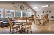 Bright communal dining area with wooden tables and chairs, sofas, large windows, and wall decorations.