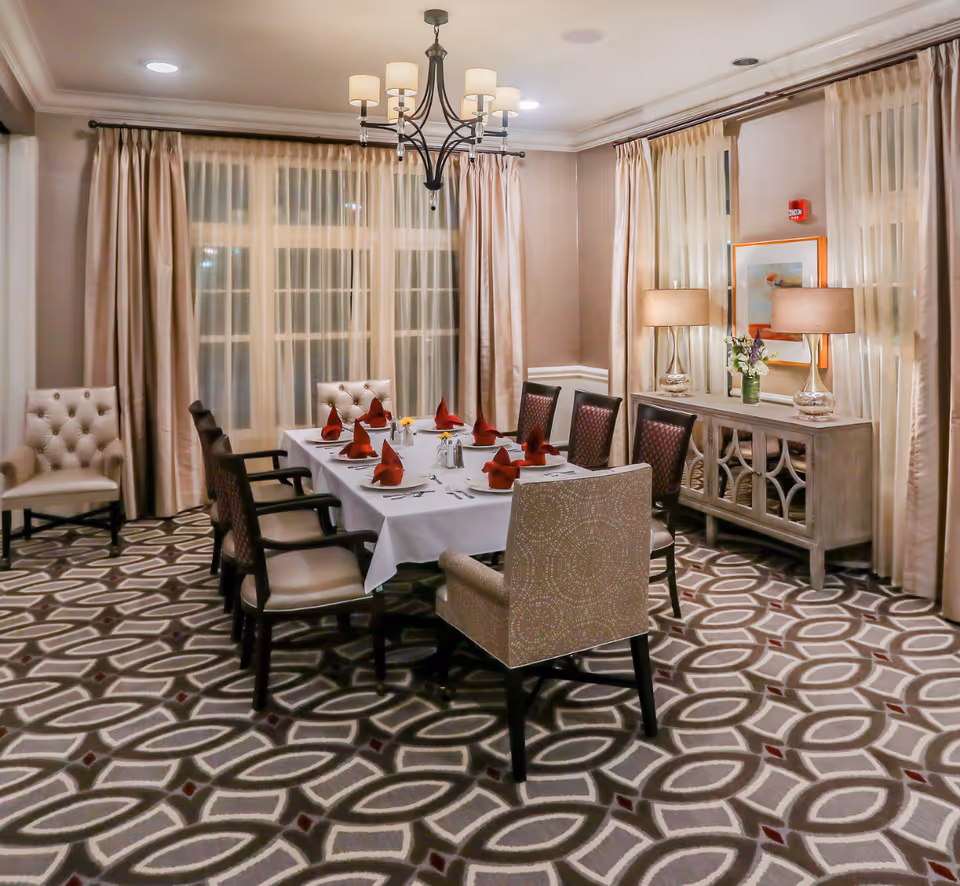 A formal dining room with a set table surrounded by upholstered chairs, a chandelier overhead, and a sideboard with lamps in front of curtained windows.