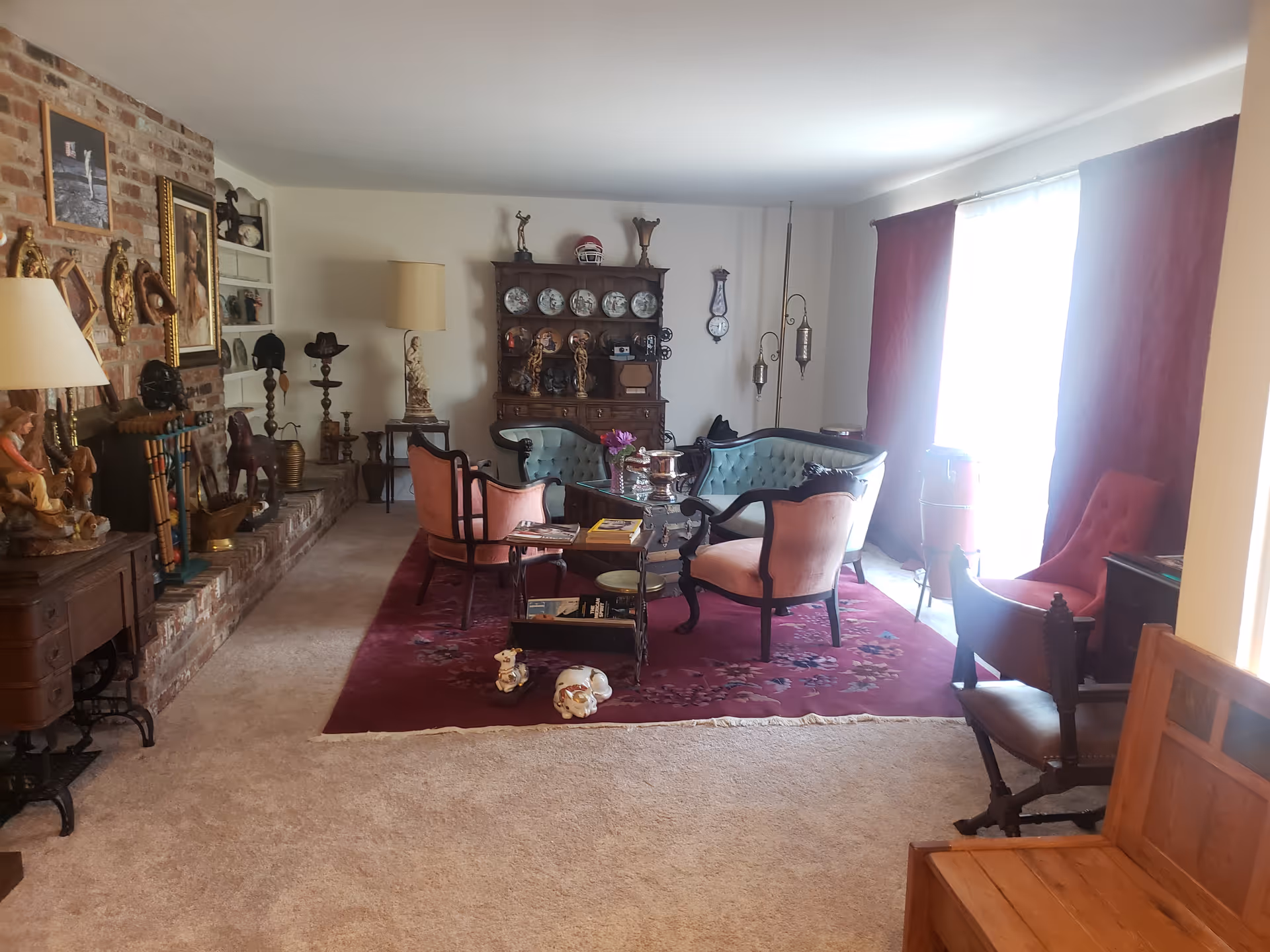 A cozy living room with vintage furniture including a tufted blue loveseat, two pink upholstered chairs, and a wooden coffee table on a red patterned rug. The room features a brick fireplace adorned with various decorative items and paintings on the wall. A wooden cabinet with plates and figurines is against the far wall. Large windows with red curtains allow natural light to brighten the space.