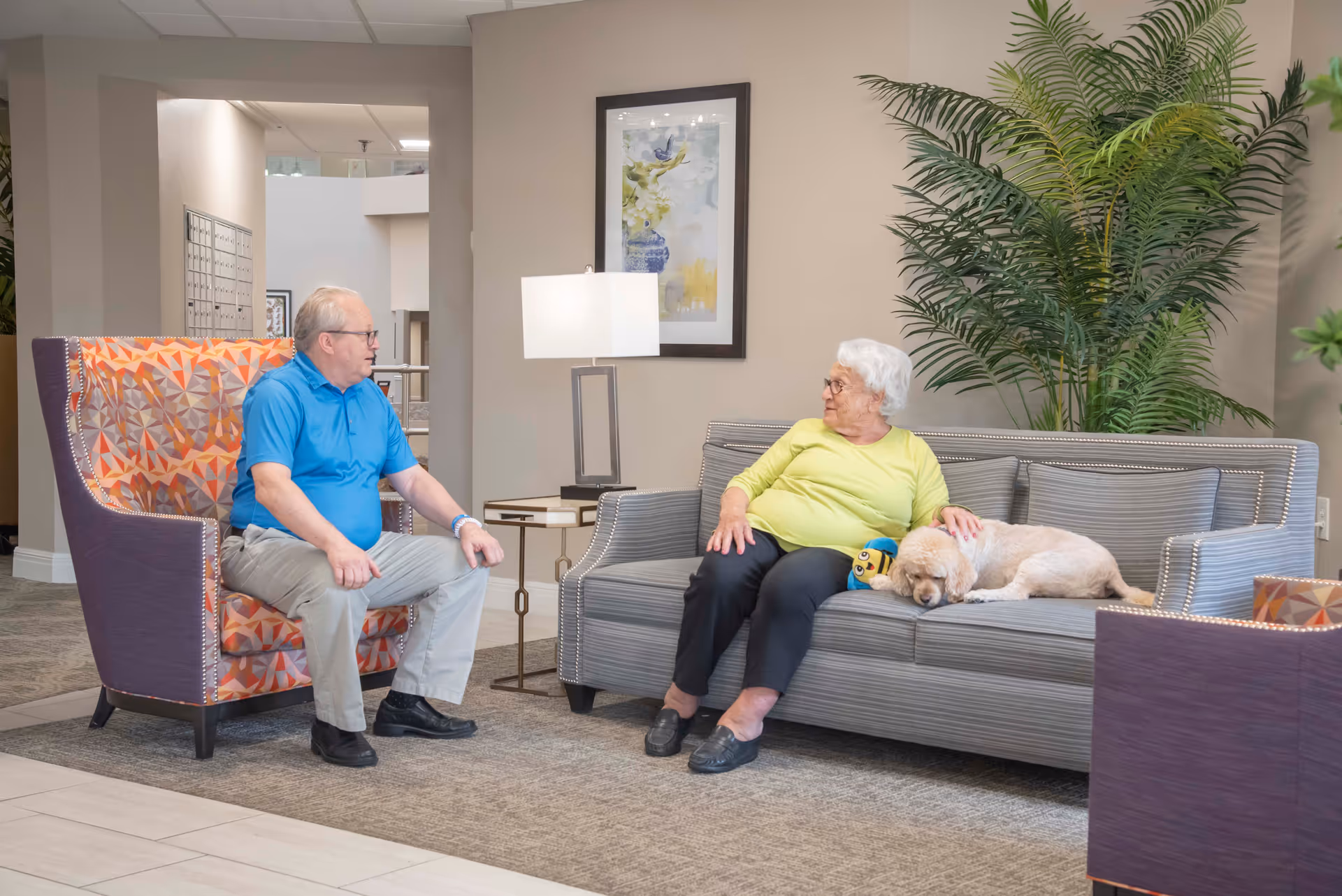 An elderly man and woman sitting in a senior living community lounge area. The man is seated in a colorful patterned armchair, and the woman is sitting on a gray sofa petting a small dog lying beside her. There is a floor lamp and framed artwork on the wall behind them, with a large green plant in the corner.