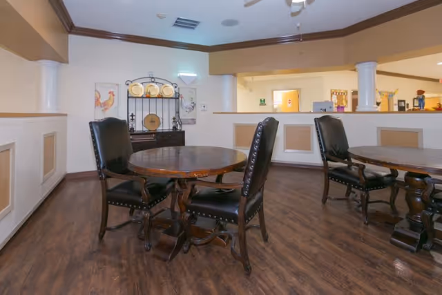 Interior view of a common area with wooden round tables and black cushioned chairs on a wooden floor. The walls are beige with decorative molding, and there is a counter with an open window to another room. Decorative plates and artwork featuring roosters are displayed on a shelf against the wall.
