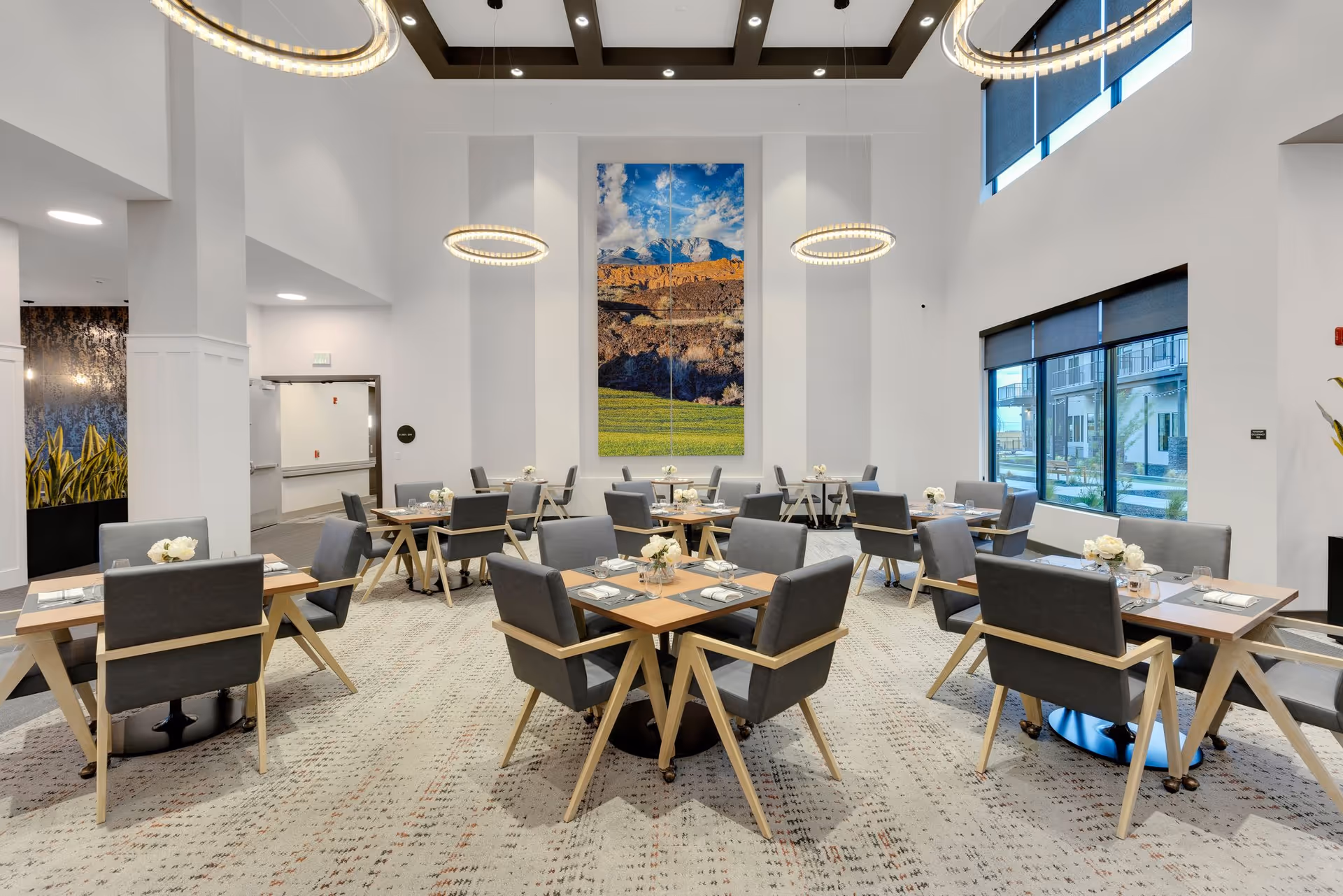 Bright modern dining room with multiple set tables and gray chairs under circular pendant lights and a large landscape artwork on the wall.
