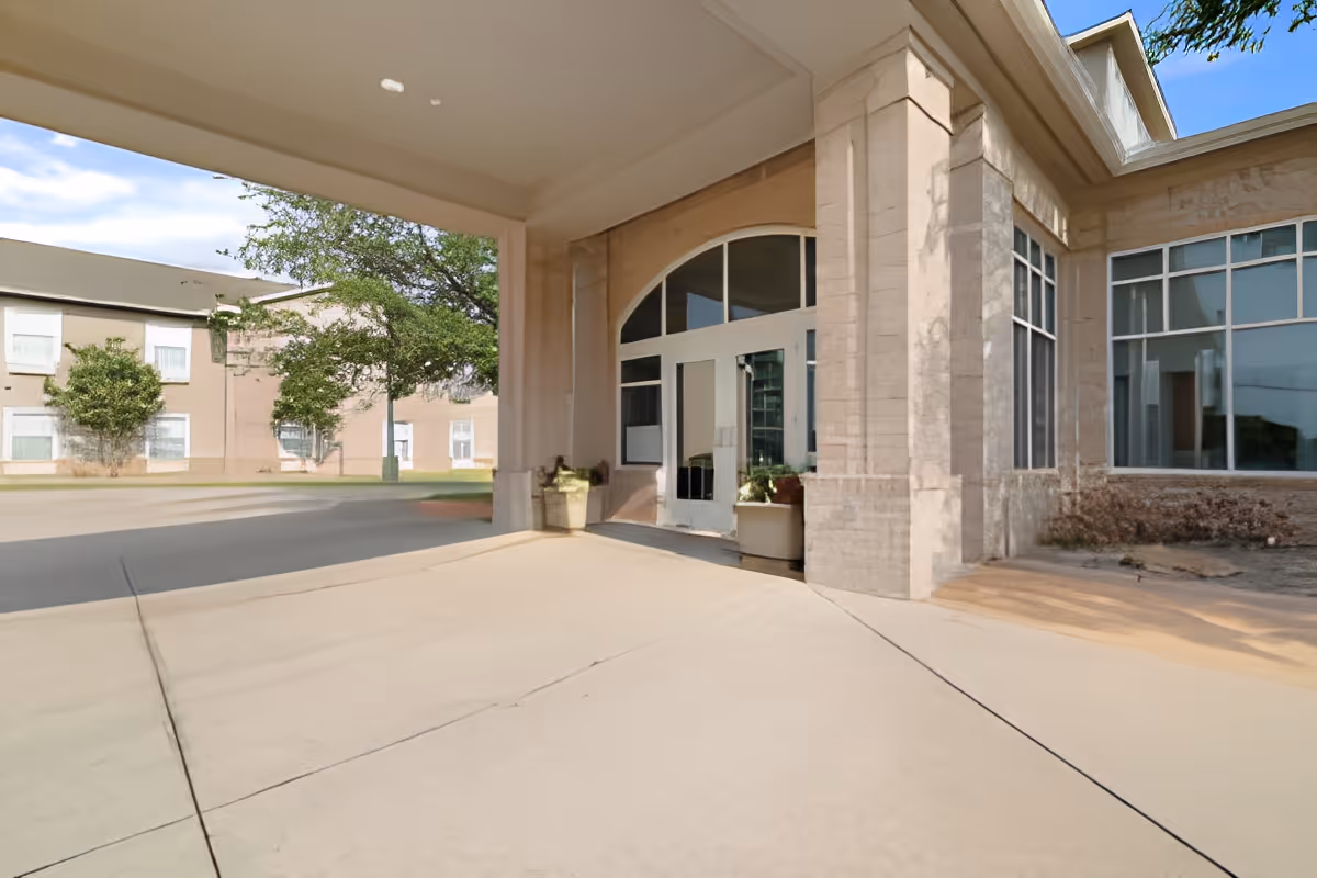 Covered entrance area of a building with large glass doors and windows, beige brick walls, and potted plants near the entrance. Trees and another building are visible in the background under a clear blue sky.