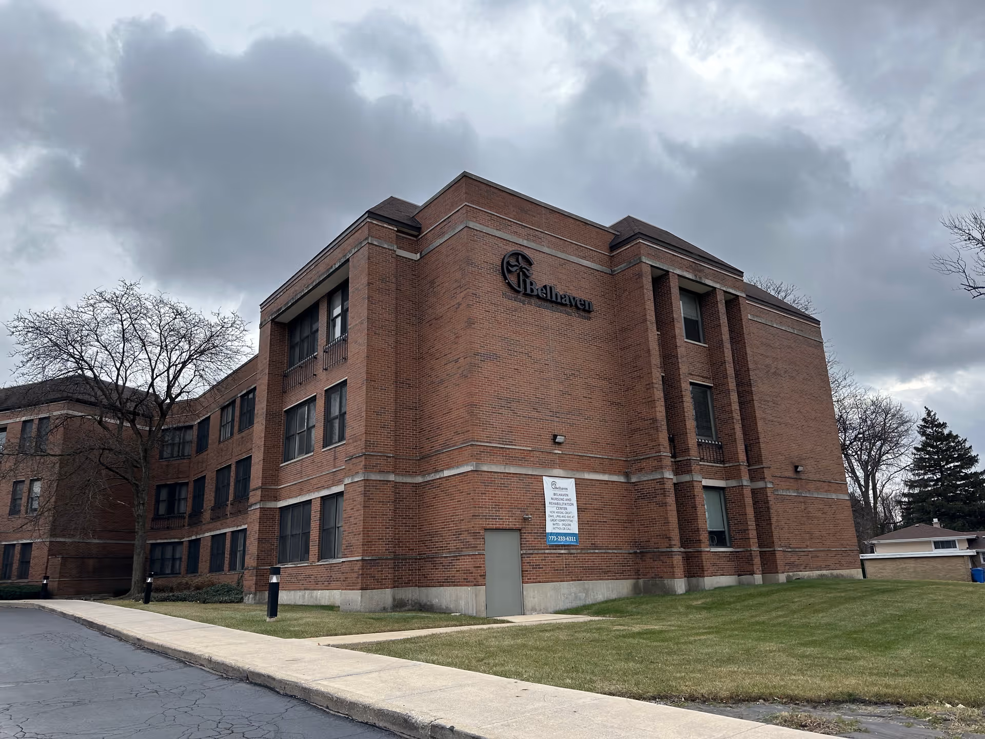 Exterior view of a three-story brick building with multiple windows, a sidewalk, and a grassy area. The building has a sign that reads 'Belhaven Nursing and Rehabilitation Center' on the wall. The sky is cloudy and overcast.