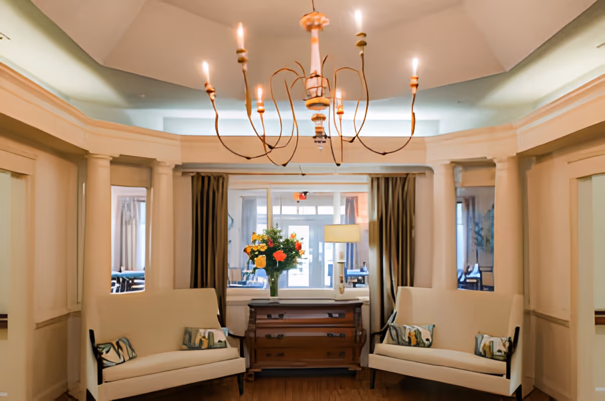 A cozy interior lounge area with two cream-colored sofas facing each other, each with decorative pillows. Between the sofas is a wooden chest of drawers with a vase of colorful flowers and a table lamp on top. The room features classical architectural details including columns and mirrors on the walls, with a large, elegant chandelier hanging from the ceiling.